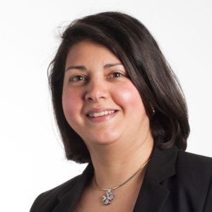 Smiling person in a black blazer with short dark hair against a white background.